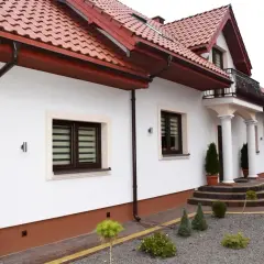 Biały dom z brązowym dachem i rynnami. Elewacja ozdobiona rustykalnym cokół. Przed domem schody i zadbany ogród.