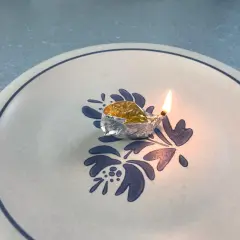 Mała lampka z folii aluminiowej, w której pali się knot zanurzony w naftcie, stoi na talerzu z niebieskim wzorem.