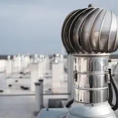 Metalowy wiatrak wentylacyjny na dachu, element systemu wentylacji hybrydowej, w tle rzędy podobnych urządzeń.