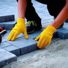 Ręce w żółtych rękawiczkach układają kostkę brukową. Obok leży gumowy młotek i miarka. Sprawdzanie układanie kostki brukowej cena.