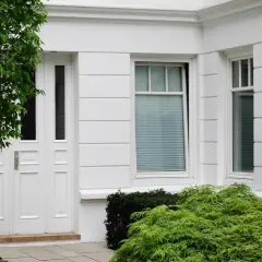 Białe drzwi i okna eleganckiego domu, otoczone bujną zielenią. Wśród roślin widać drzewo o gęstych liściach i krzewy, tworzące naturalne bonie.