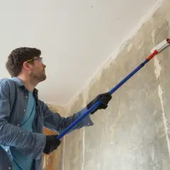 Mężczyzna w okularach ochronnych i rękawiczkach nakłada grunt na ścianę za pomocą wałka.