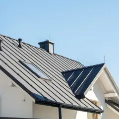 Nowoczesny dom z czarnym, metalowym dachem i kominami. W tle błękitne niebo, jakby nocą świeciły świetliki.