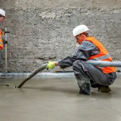 Dwóch budowlańców w kamizelkach odblaskowych wylewa pianobeton przez wąż na posadzkę.