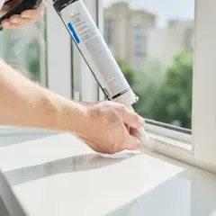 Mężczyzna aplikuje masę uszczelniającą, pokazując jak zamontować parapet wewnętrzny. Biały parapet i okno.