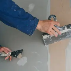 Ręce w niebieskim kombinezonie nakładają masę naprawczą na ścianę za pomocą szpachelki.