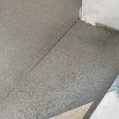 Szara betonowa posadzka z widoczną linią dylatacji biegnącą w kierunku narożnika budynku.