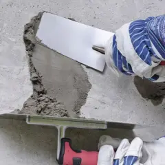 Naprawa betonu na zewnątrz. Ręce w rękawicach nakładają zaprawę na uszkodzony fragment ściany za pomocą kielni.
