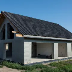 Dom w stanie surowym otwartym, z dachem pokrytym czarną dachówką. Ściany z bloczków betonowych, bez okien i drzwi.