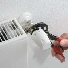 Dłoń z kombinerkami dokręca termostat grzejnika. Regulacja temperatury w domu to klucz do komfortu.