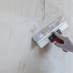 Ręka w rękawicy wykonuje szpachlowanie ścian, wygładzając nierówności.