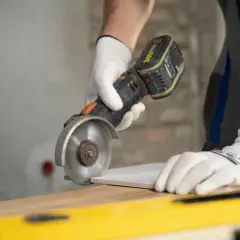 Ręce w białych rękawiczkach precyzyjnie wykonują fazowanie płytki ceramicznej za pomocą szlifierki kątowej.