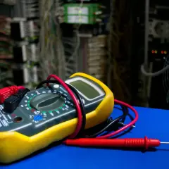 Żółty multimetr z czerwonymi i czarnymi przewodami na tle szafy elektrycznej.