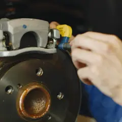 Mechanik dokonuje wymiany linki hamulca ręcznego, pracując przy zacisku i tarczy hamulcowej.
