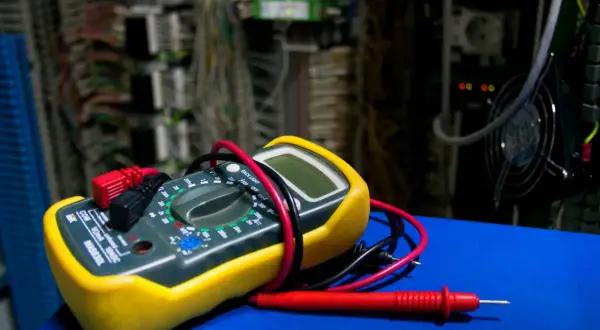 Żółty multimetr z czerwonymi i czarnymi przewodami na tle szafy elektrycznej.