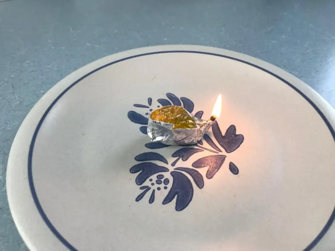 Mała lampka z folii aluminiowej, w której pali się knot zanurzony w naftcie, stoi na talerzu z niebieskim wzorem.