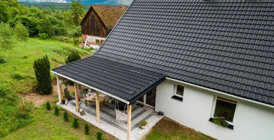 Nowy, ciemnoszary dach z blachodachówki nad tarasem i domem, otoczony zielenią.