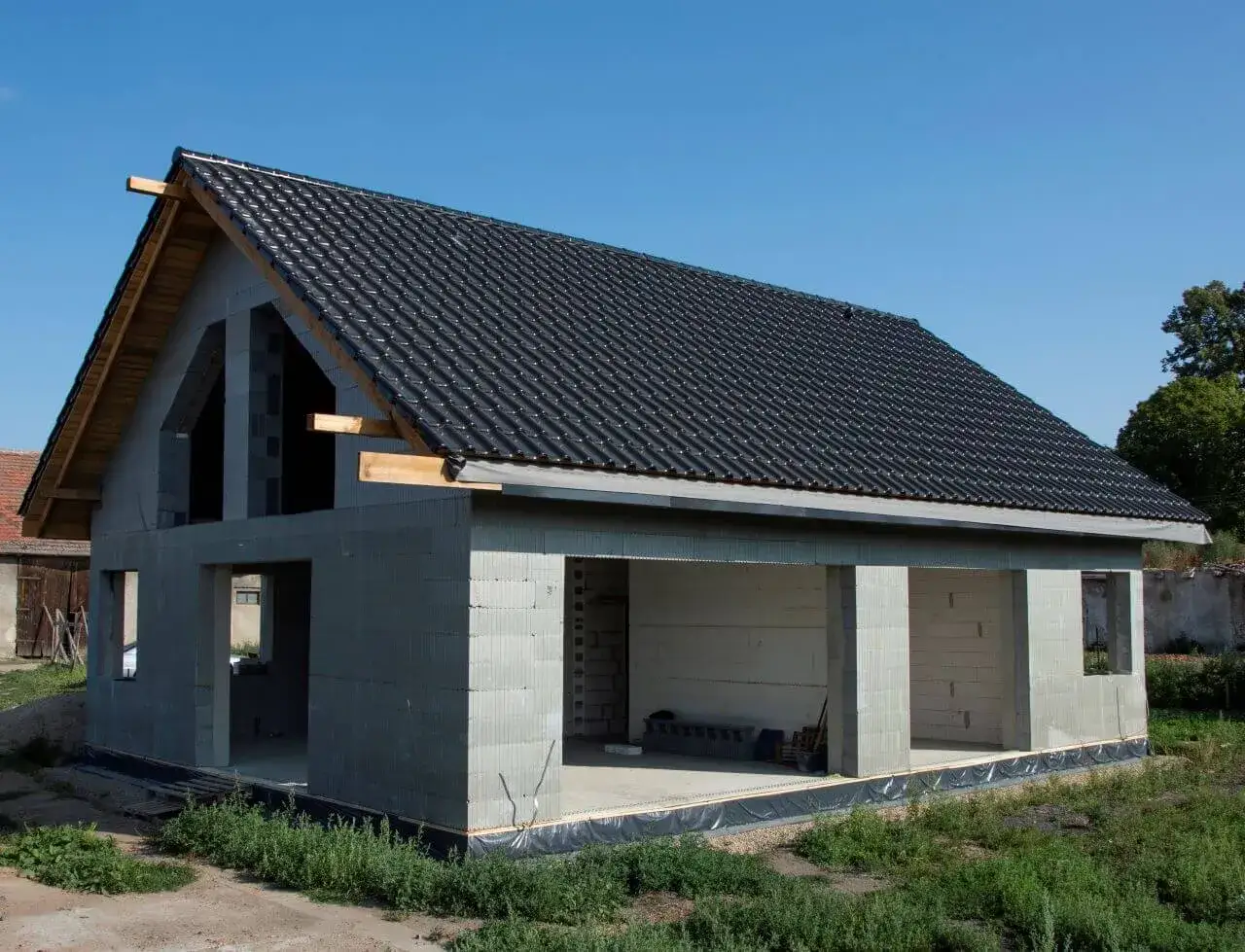 Dom w stanie surowym otwartym, z dachem pokrytym czarną dachówką. Widoczne betonowe ściany i otwory okienne oraz drzwiowe.