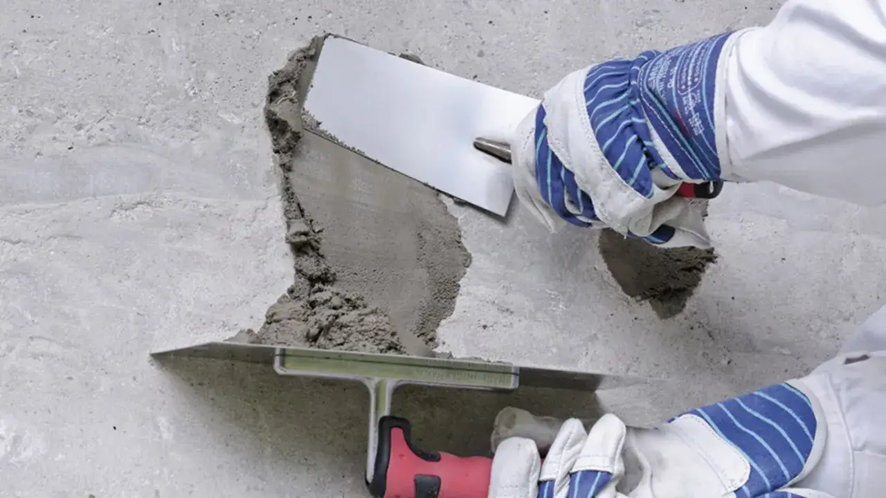 Naprawa betonu na zewnątrz: ręce w rękawicach nakładają zaprawę kielnią na uszkodzoną powierzchnię.