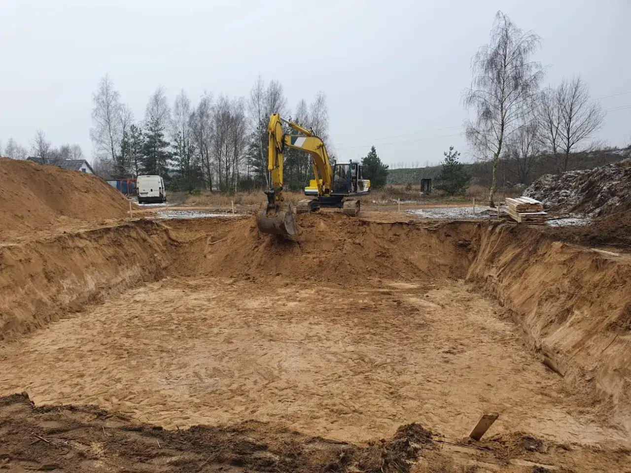 Koparka pracuje przy wykopy pod fundamenty. Widać głęboki dół, usypiska ziemi i materiały budowlane.