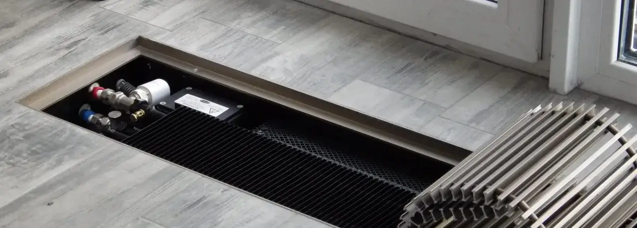 Ukryty w podłodze konwektor kanałowy z przyłączami hydraulicznymi i czarną kratką. Obok leży metalowa kratka maskująca.