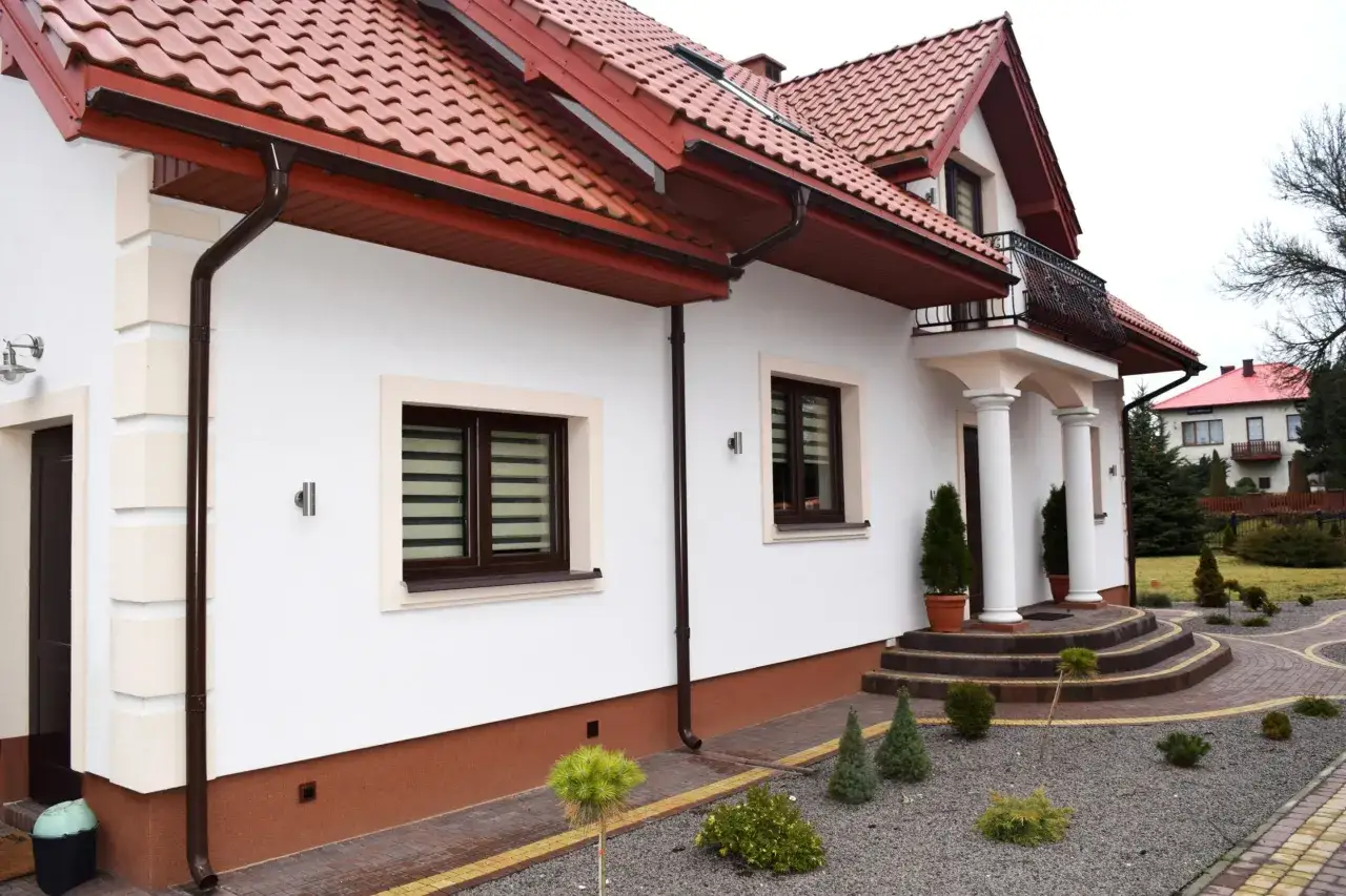 Biały dom z brązowym dachem i rynnami. Widać schody prowadzące do wejścia z kolumnami. Na pierwszym planie ozdobny cokół i kamyczki.