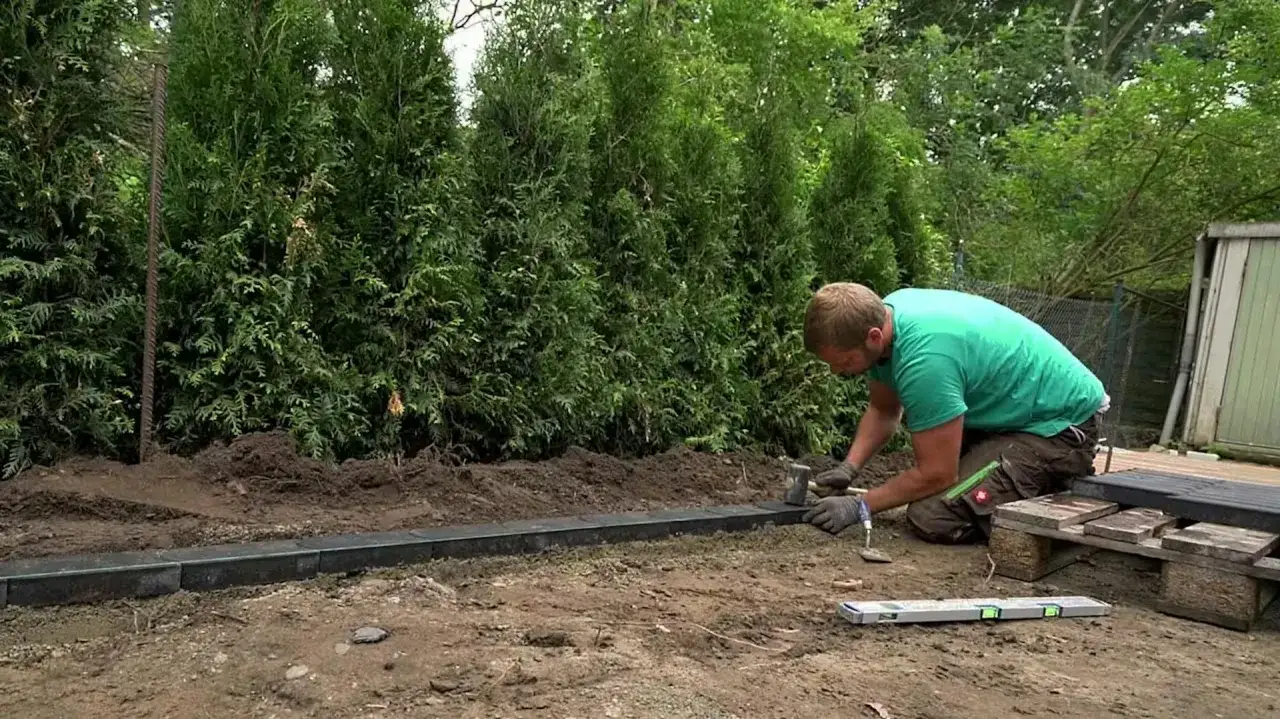 Mężczyzna klęczy i układa krawężniki w ogrodzie, używając młotka i poziomicy.
