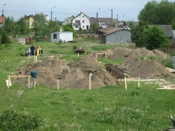 Prace budowlane w toku: kopanie wykopy pod fundamenty, ziemia usypana w kopce. W tle domy i pracownicy.